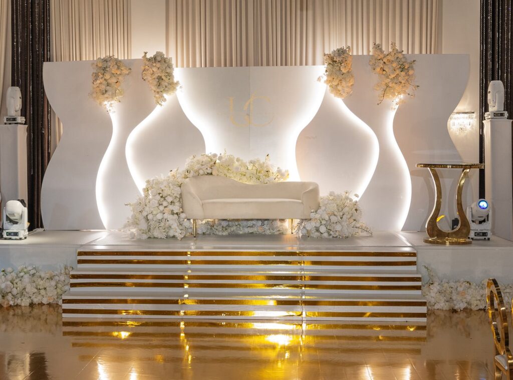Sculptural white and gold wedding stage with illuminated curved panels, floral installations, and mirrored gold steps — a photogenic luxury event backdrop.