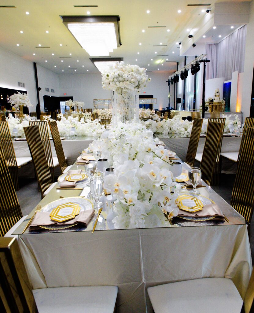 Elegant long wedding tablescape with cascading white florals, gold chargers, crystal candle holders, and gold chairs, designed by Events D’Lux for a luxury white-and-gold reception.