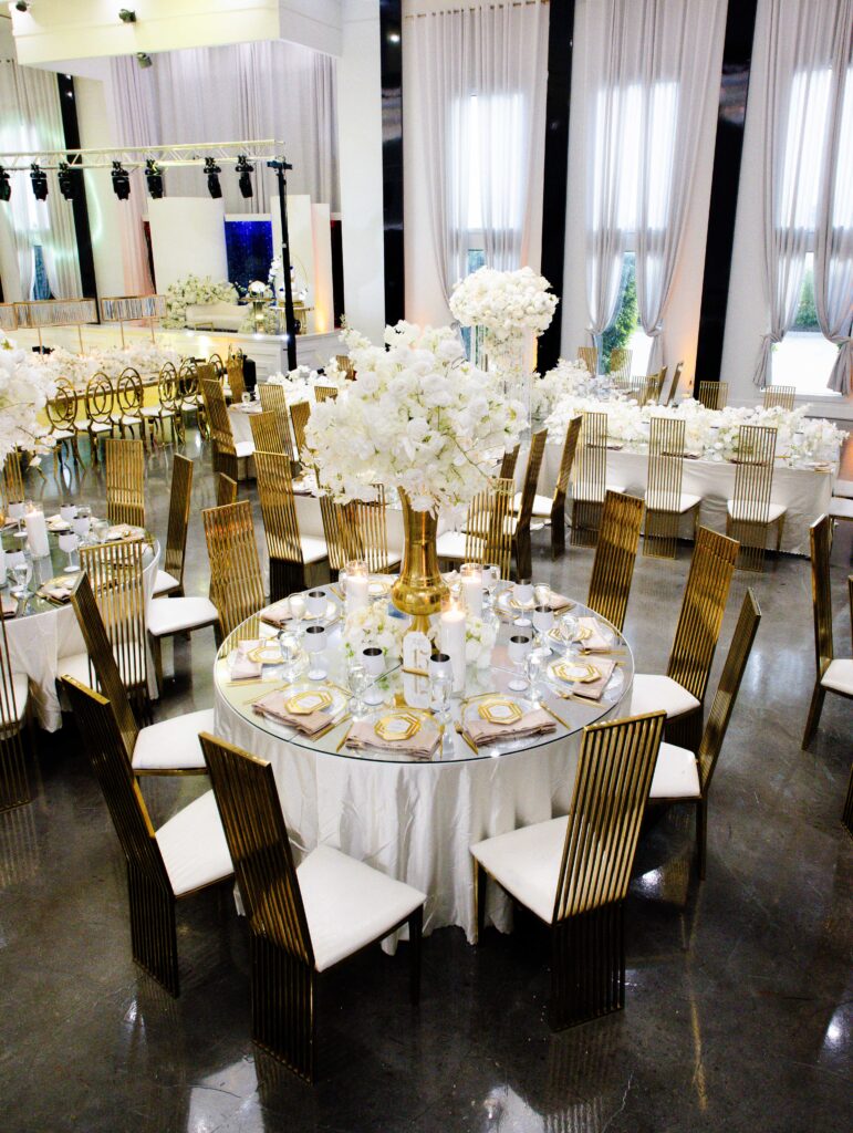 Round wedding reception table featuring a tall white floral centerpiece on a gold vase, gold-rimmed dinnerware, and gold chairs in a modern luxury ballroom.