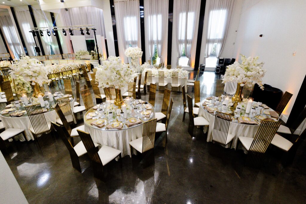 Luxury wedding reception featuring multiple round tables with tall white floral centerpieces, gold pedestal vases, and gold chairs in an airy, elegant ballroom.