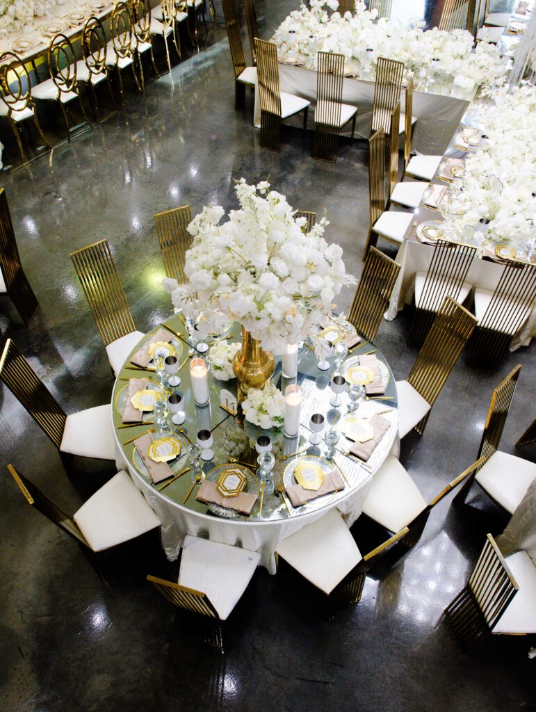 Aerial shot of a round wedding tablescape with lush white florals, gold charger plates, crystal candle holders, and gold chairs for a sophisticated reception design.