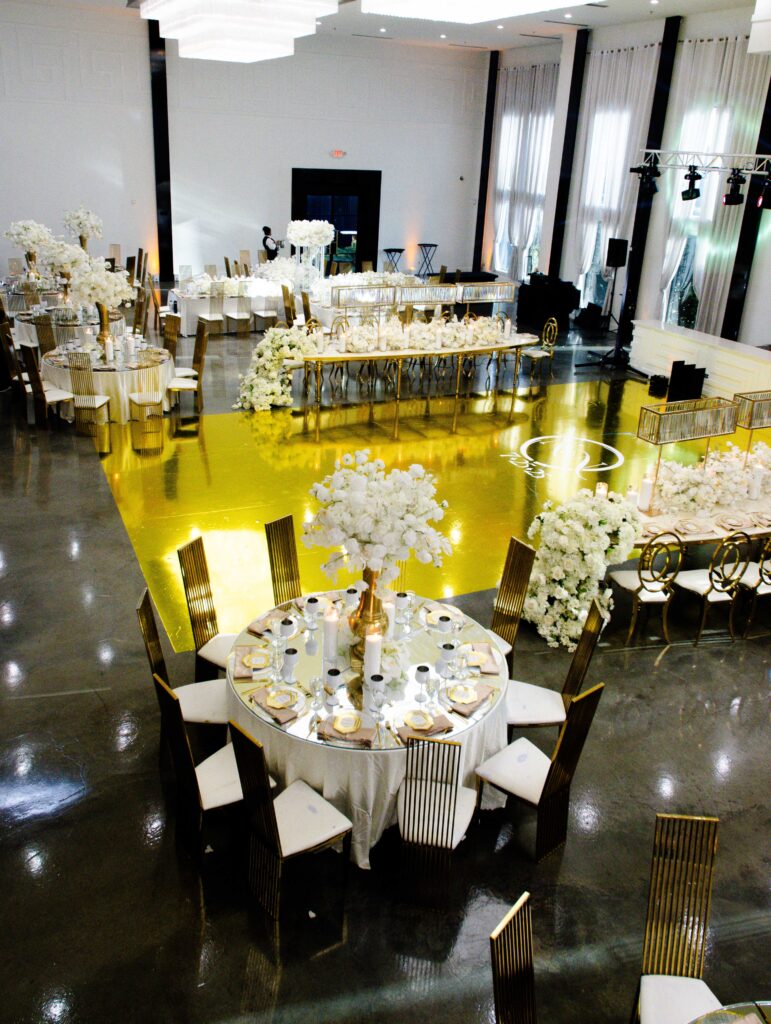 Luxury wedding reception with a custom gold mirrored dance floor, round and long tables featuring lush white floral centerpieces, gold chairs, mirrored tabletops, and elevated floral runners—an elegant white-and-gold design by Events D’Lux.