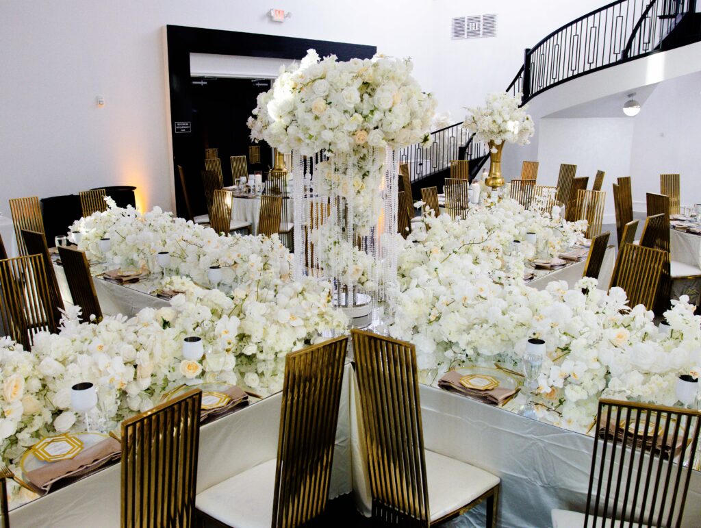 Cluster of tall white floral wedding centerpieces with crystal accents, surrounded by gold modern chairs and luxe table settings at a high-end reception by Events D’Lux.