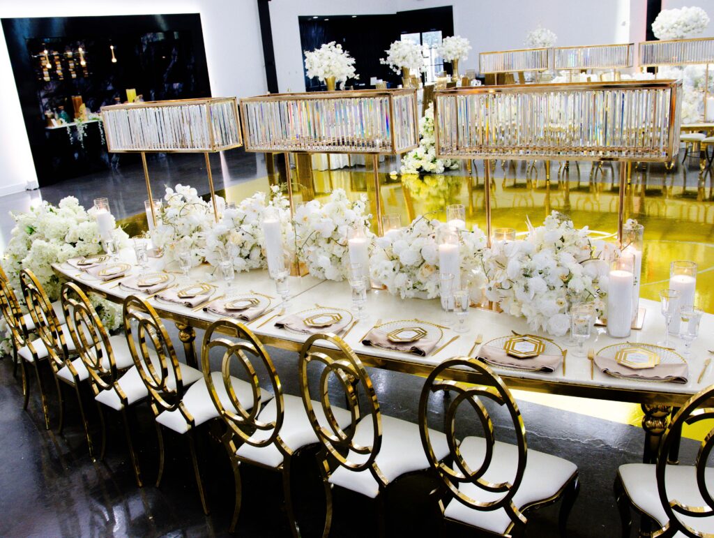 Opulent long wedding tablescape featuring gold circular-back chairs, white florals, mirrored candle holders, and gold charger plates for a sophisticated luxury reception.