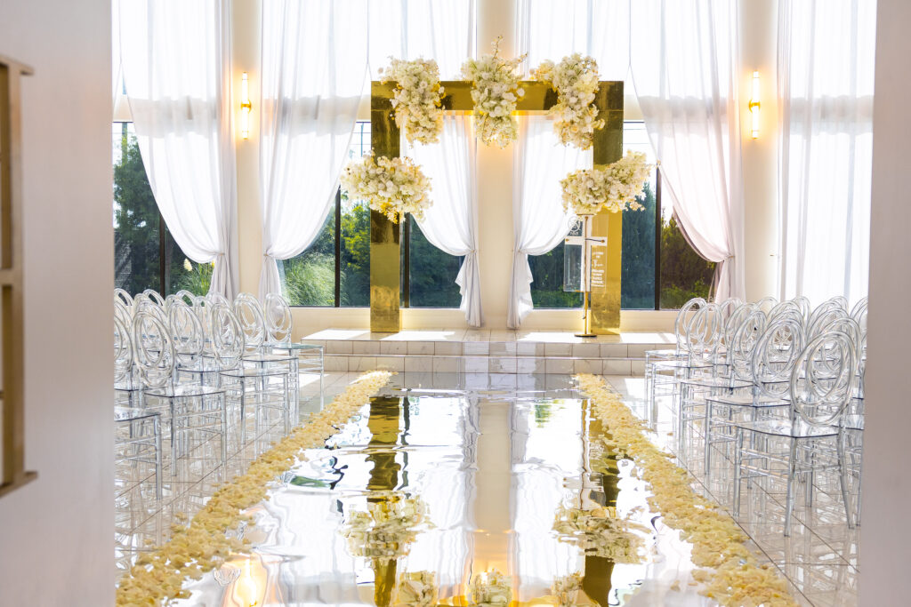 Luxury wedding ceremony setup featuring a 3D gold mirror square arbor with lush white florals and a mirrored aisle, designed by Events D’Lux at The Hamptons Event Venue chapel.