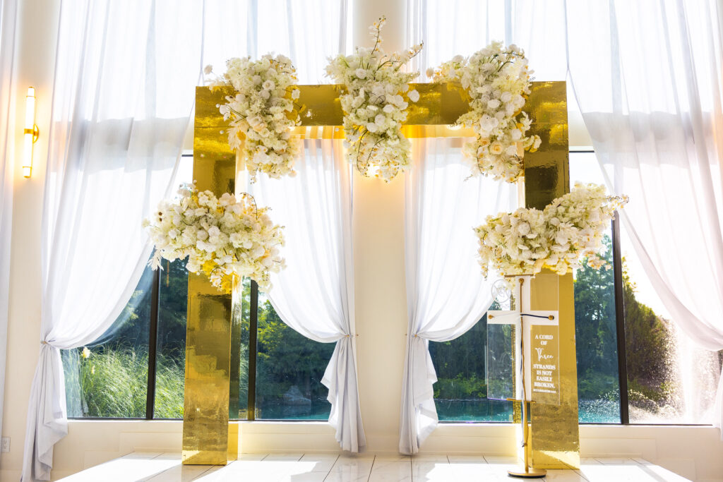 Modern gold square wedding arbor adorned with cascading white floral arrangements, creating an elegant ceremony backdrop designed by Events D’Lux.